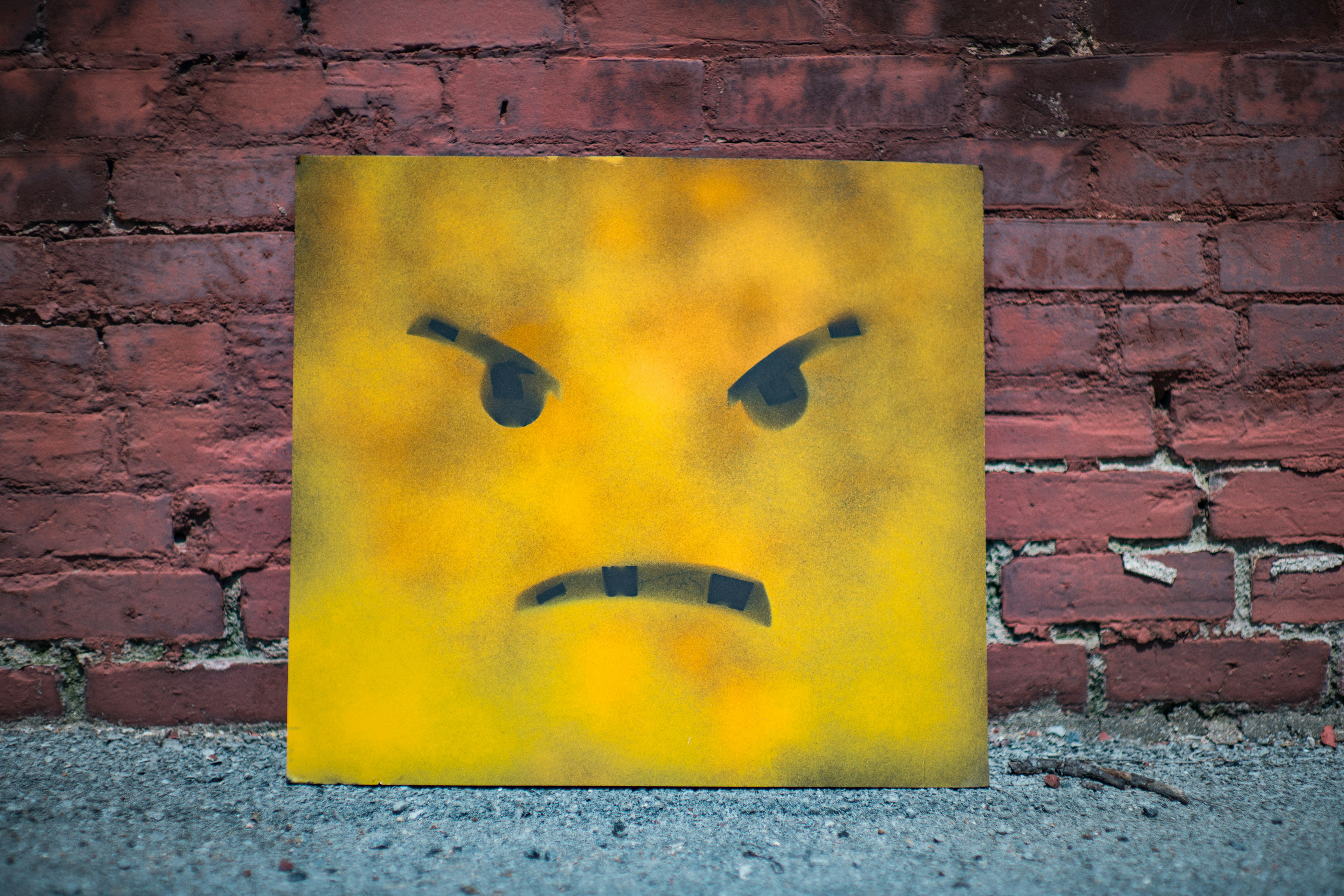 A graffiti-style angry emoji face on a yellow board against a brick wall